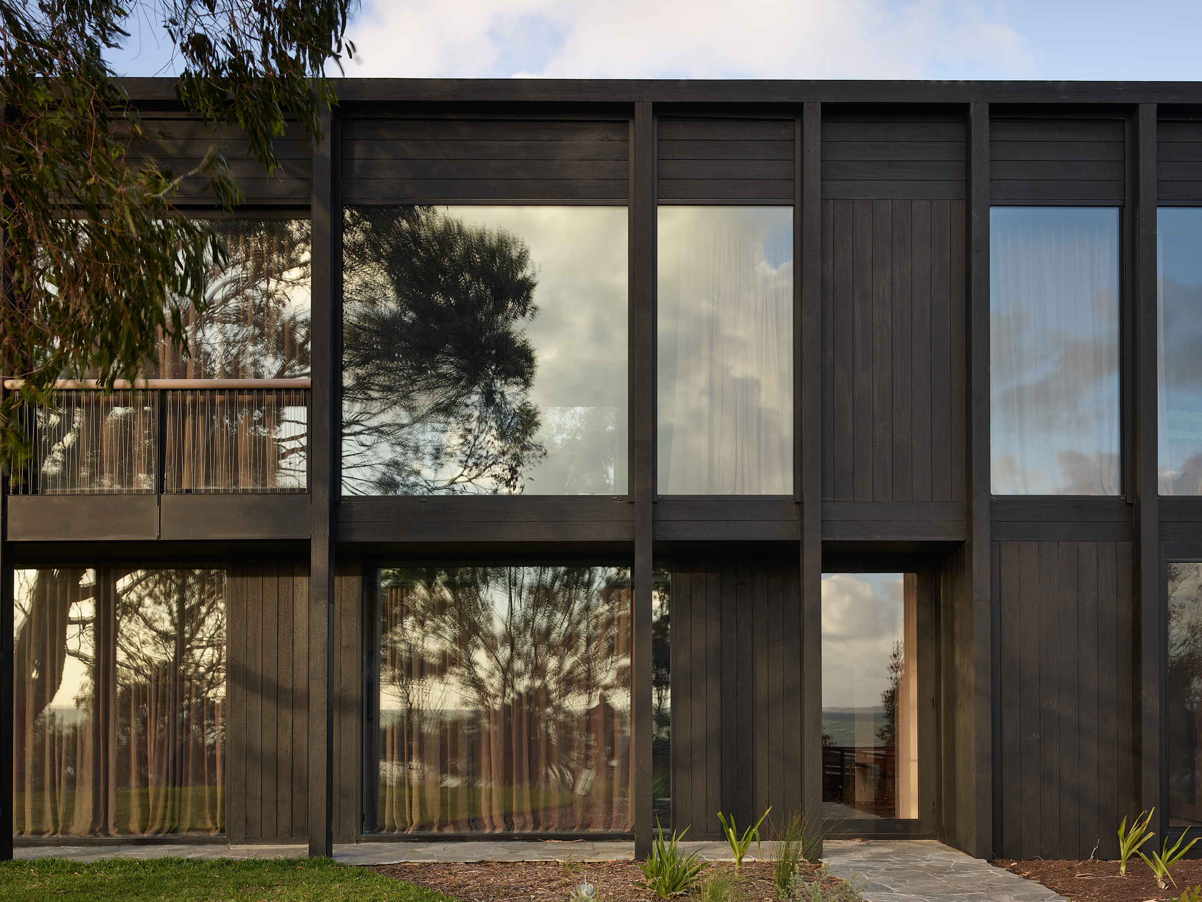 The facade of a two storey building with black timber cladding, and large glass windows that reflect the sky.